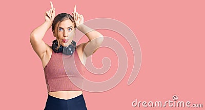 Beautiful caucasian young woman wearing gym clothes and using headphones doing funny gesture with finger over head as bull horns Stock Photo
