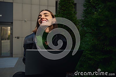 Beautiful delighted freelancer confident young woman smiles , looking up with closed eyes of pleasure , working remotely outdoor Stock Photo