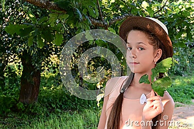 beautiful cheerful young girl in hat stands among green foliage and holds leaf in her hand. Proximity to nature. Stock Photo