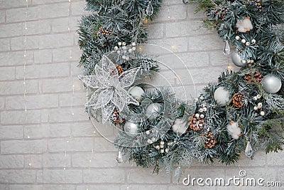 Beautiful Christmas wreath of fir branches, cones, white baubles, flowers, berries and icycles on brick wall with garland lights. Stock Photo