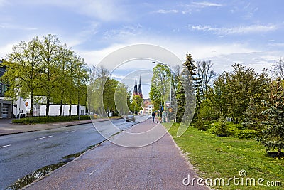 Beautiful cityscape view on bright day. Man on scooter and vehicle on road with green trees on sides and church towers on blue sky Editorial Stock Photo