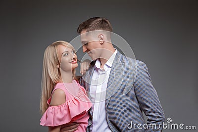 Beautiful couple is looking at each other and smiling while standing straight on gray background. Stock Photo