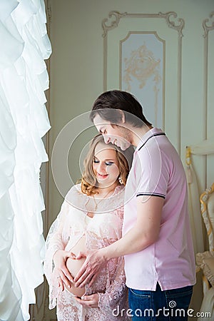 A beautiful couple, a pregnant young woman and a man stand near a window, Make a heart from the hands on the stomach. Stock Photo