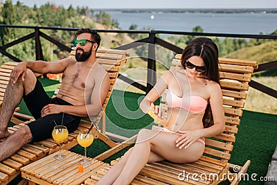 Beautiful couple sit on sunbeds and chilling. Girl put some cream protection on skin and rub it. Guy is calm. He looks Stock Photo