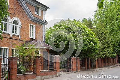 Beautiful courtyard with a brick red house, forged gates and lush flowering chestnut. Pretty country house patio, blooming green Stock Photo