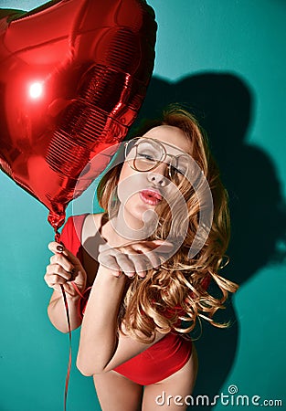 Beautiful curly hair young woman in clear aviator glasses hold big red star balloon blow kiss sign with lips on mint background Stock Photo