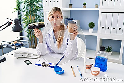 Beautiful dentist woman holding denture and toothbrush making fish face with mouth and squinting eyes, crazy and comical Stock Photo