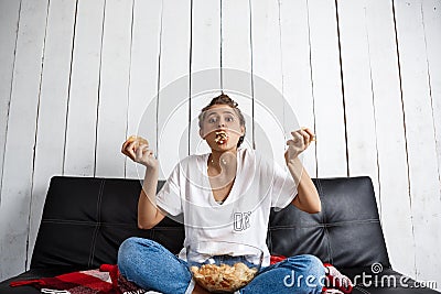 Beautiful domestic girl eating chips, watching tv, sitting at sofa. Stock Photo