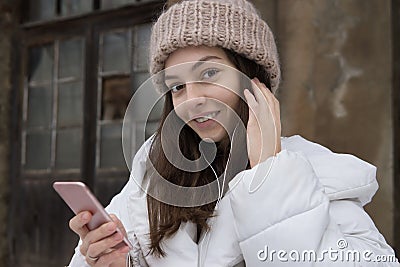 Beautiful European girl in a white jacket and knitted hat listening to music with headphones walking around the city on Stock Photo