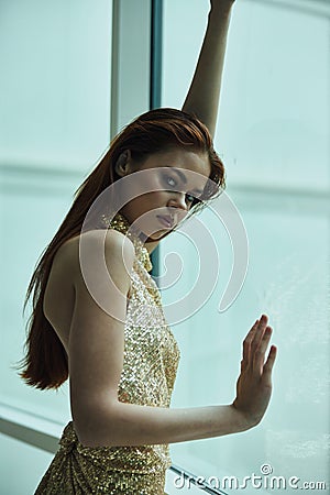 Beautiful fashion woman in stylish expensive gold sequined dress poses looking out the window at the city in a hotel Stock Photo