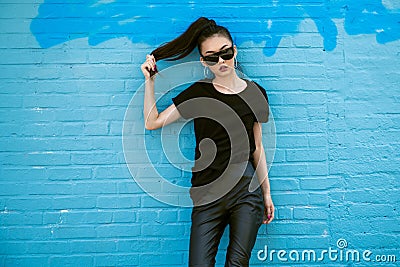 Beautiful fashionable asian girl wearing sunglasses and black clothes outfit posing in front of blue wall. Stock Photo