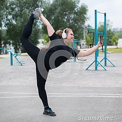 Beautiful fat young woman in headphones makes a figure skater and takes a selfie. A chubby smiling girl listens to music Stock Photo