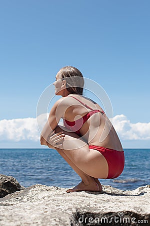 Beautiful female person in red bikini sits squatting on rock Stock Photo