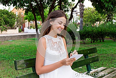 Beautiful female student is writing her ideas and thoughts into the notebook sitting on the bench in the park Stock Photo