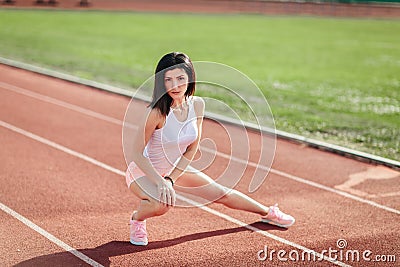 Beautiful fitness brunette girl in sportswear and sneakers does a stretching exercise on the running track at the stadium outdoors Stock Photo