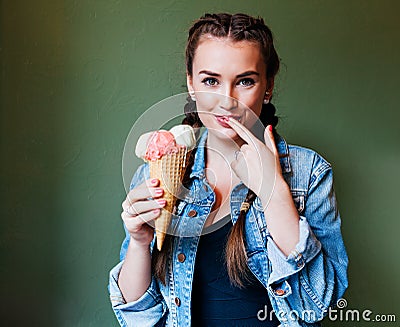 Beautiful girl with braids sitting in a cafe and eating huge multi-colored ice cream in a waffle cone. Licking finger Stock Photo