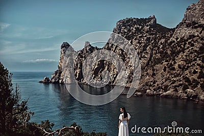 A beautiful girl, a bride, in a white dress stands on a cliff against the background of the sea and mountains. Stock Photo