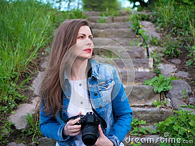 Beautiful girl in a cotton jacket sits on vintage stone steps and taking photos with a professional camera in windy weather Stock Photo