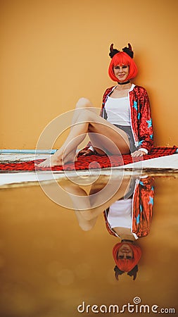 Beautiful girl devil sitting on an orange background near the puddle with his reflection. In anticipation of Halloween Stock Photo