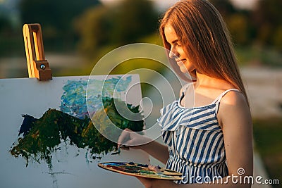 Beautiful girl draws a picture in the park using a palette with paints and a spatula. Easel and canvas with a picture. Summer is a Stock Photo