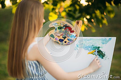Beautiful girl draws a picture in the park using a palette with paints and a spatula. Easel and canvas with a picture. Summer is a Stock Photo