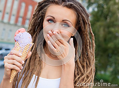 Beautiful girl with dreadlocks eating colorful ice cream and covers her mouth a palm on a warm summer night in the street. Outdoor Stock Photo