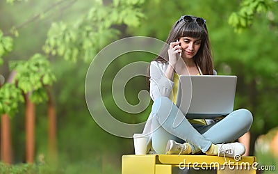 Cute girl freelancer is working on a laptop in the park and talking on the phone. Remote work concept Stock Photo