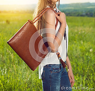 Beautiful girl holding brown leather hand bag outdoors on the sunny meadow at sunset time. Girl wearing fashionable white t-shirt Stock Photo