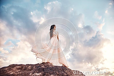 Beautiful girl in a light summer dress beige walks in the mountains. Light dress flutters in the wind, blue summer sky. Fabulous Stock Photo