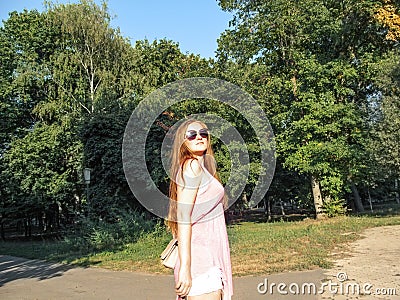 Beautiful girl with long golden hair in sunglasses looks at the sun. Young woman in pink clothes walking in a park on a clear Stock Photo