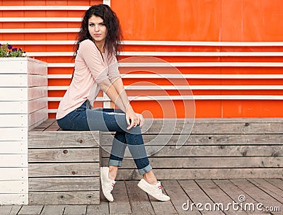 Beautiful girl with long hair brunette in jeans sits near wall of orange old white wooden planks Stock Photo