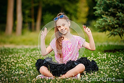 Beautiful girl with a long tail of hair in a full skirt bundle sits on the grass with her hands up and smiling Stock Photo