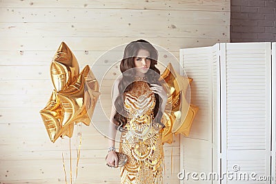 Beautiful girl with long wavy shiny hair. Brunette woman with elegant hairstyle in golden dress over party star balloons. Party Stock Photo