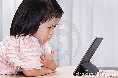 Beautiful girl lying down and playing game on tablet on wooden table. Child watching cartoon with touchpad. Kid using technology Stock Photo