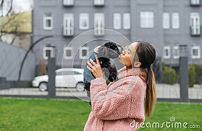 Beautiful girl owner in pink coat holds black curly puppy in her hands and kisses, doggy looks into the camera. Walk with your pet Stock Photo