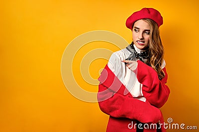 A beautiful girl in a red coat and beret stands on a yellow background with horror, looks away pointing finger there Stock Photo