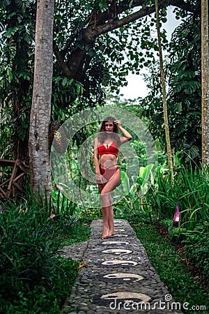 Beautiful girl in red swimsuit posing in tropical location with green trees. Young sports model in bikini with perfect Stock Photo