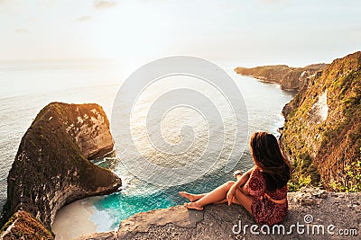 Beautiful girl sitting on a high rock and looking out to sea, rear view. Girl sits on the edge of cliff. The girl of the sea. Stock Photo