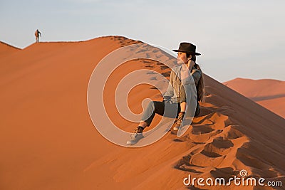 A beautiful girl tourist in a cowboy hat sits on the top of a dune in the Namib desert. Sossuflei, Namibia, Africa. Stock Photo