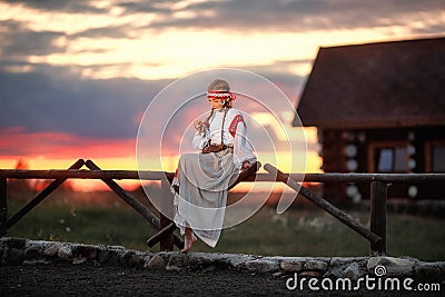 A beautiful girl in a traditional Russian dress is sitting on a fence against the sunset Stock Photo