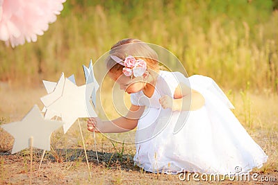 Beautiful girl in white long dress puts paper star on ground. Child on background of nature. Stock Photo