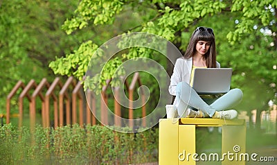 Beautiful girl works sitting with a laptop in a park on a background of green trees. Freelance work in a recreation area Stock Photo