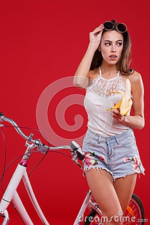 A girl raises her glasses and looks to the side with a banana in her hand on a red background Stock Photo