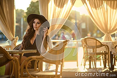 Beautiful glam tattooed brunette in little black dress and trendy fedora hat sitting in the nice open air summer restaurant Stock Photo