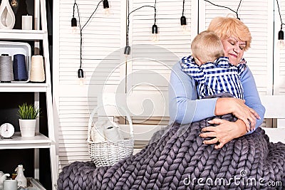 A beautiful grandmother and grandson are sitting on the sofa under a knitted blanket merino wool and tender embrace. A happy famil Stock Photo