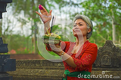 Beautiful and happy Asian Indonesian woman dressed in traditional Balinese religious custom holding incense stick and flowers Stock Photo