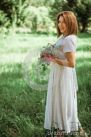 A beautiful happy girl in a white dress holds a bouquet of wild flowers in her hands. Portrait of a slender girl with a bouquet of Stock Photo