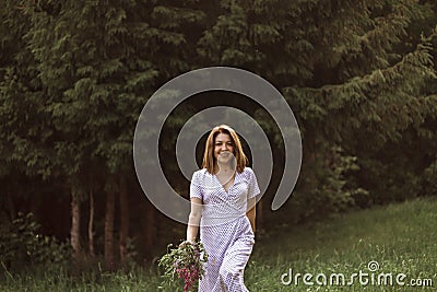 A beautiful happy girl in a white dress holds a bouquet of wild flowers in her hands. Portrait of a slender girl with a bouquet of Stock Photo