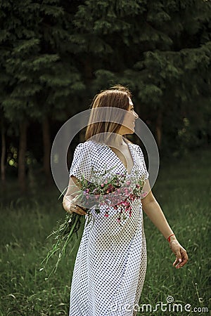 A beautiful happy girl in a white dress holds a bouquet of wild flowers in her hands and turns around. Portrait of a slender girl Stock Photo