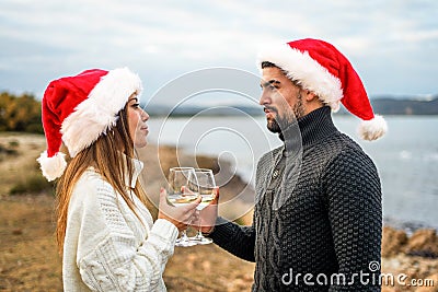 Beautiful heterosexual couple in love toasting outdoor wearing Santa Claus hat looking into each other`s eyes holding white wine Stock Photo